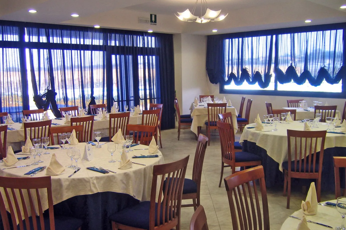 Sala da pranzo con tavoli rotondi apparecchiati e ampie vetrate con tende blu