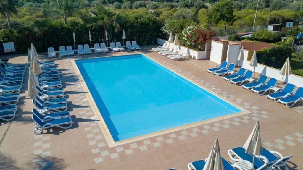 Piscina rettangolare circondata da lettini blu e ombrelloni, con vegetazione e olivi sullo sfondo