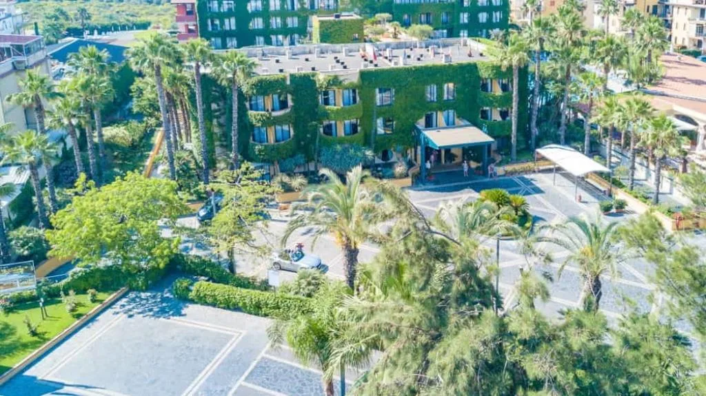 Vista aerea dell'hotel con facciata verde, ampio piazzale e giardino di palme