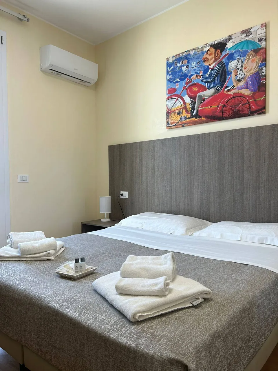 Camera matrimoniale con testiera in legno, quadro colorato, aria condizionata e asciugamani sul letto