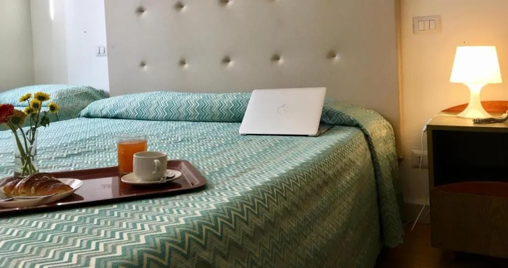 Camera con letto matrimoniale, copriletto verde a zigzag, laptop e comodino illuminato