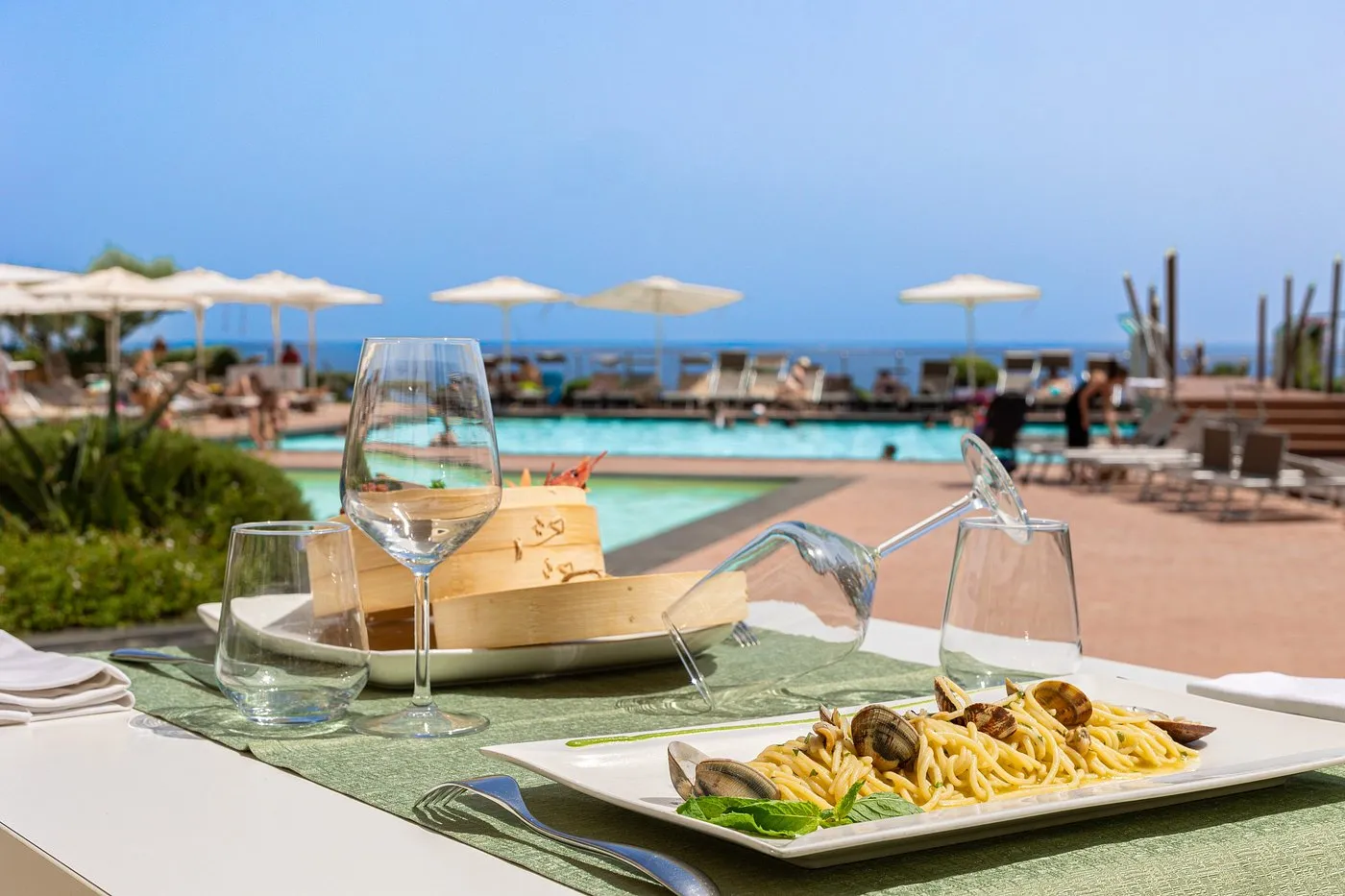 Tavolo apparecchiato a bordo piscina con piatto di spaghetti alle vongole e vista mare