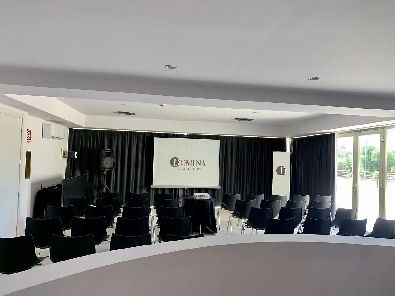 Sala meeting con sedie nere, schermo per proiezioni e logo Domina