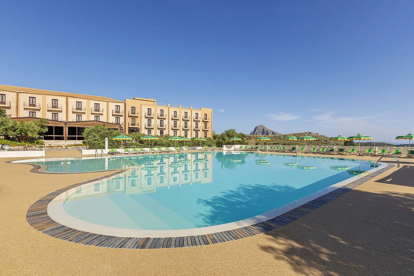 Ampia piscina esterna a forma sinuosa con lettini verdi accanto all'edificio dell'hotel e montagna sullo sfondo