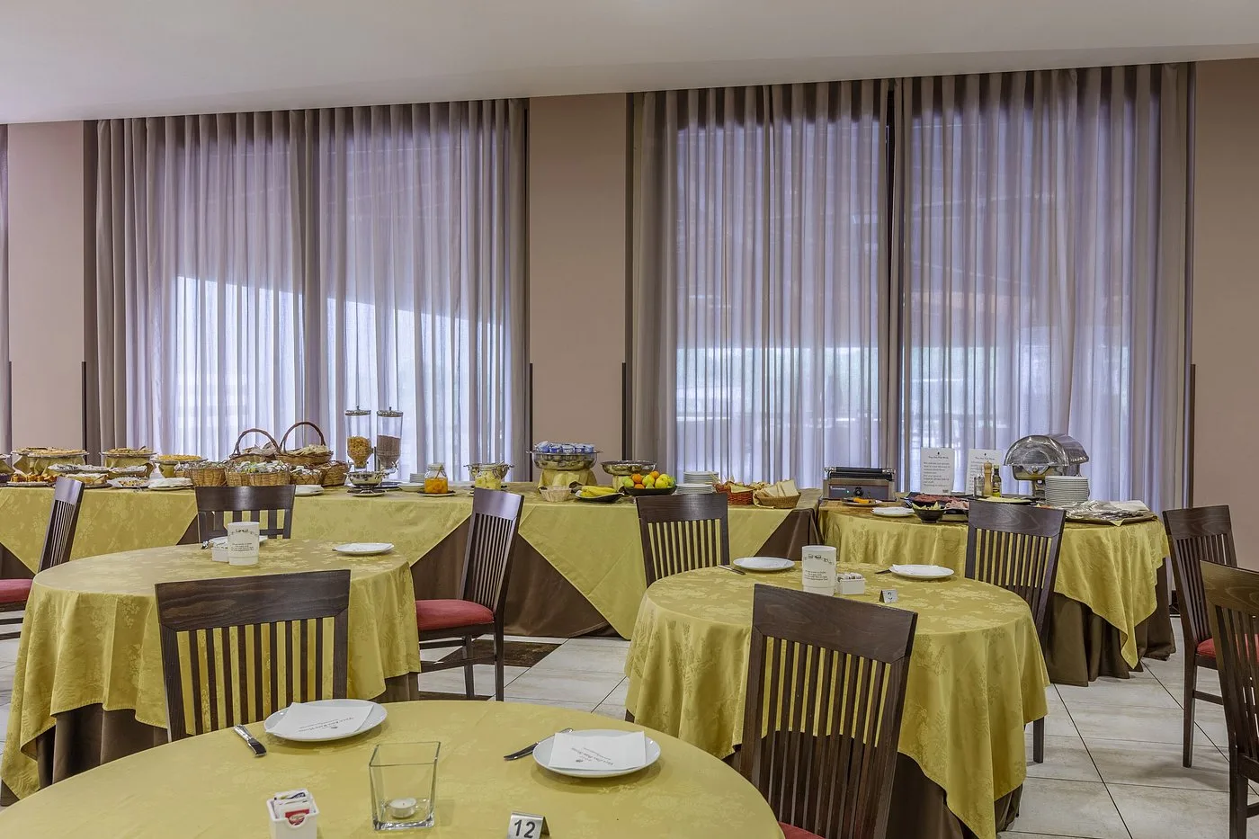 Sala ristorante con buffet, tavoli rotondi con tovaglie gialle e ampie finestre con tende