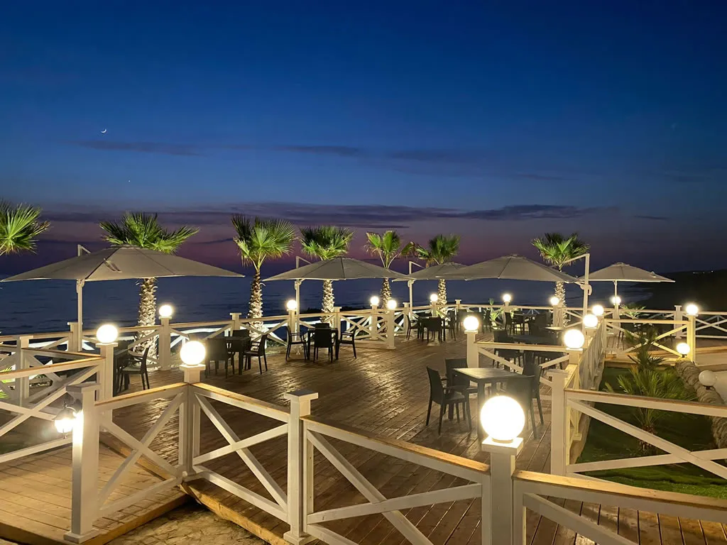 Terrazza in legno con ombrelloni, palme e luci sferiche al crepuscolo sul mare