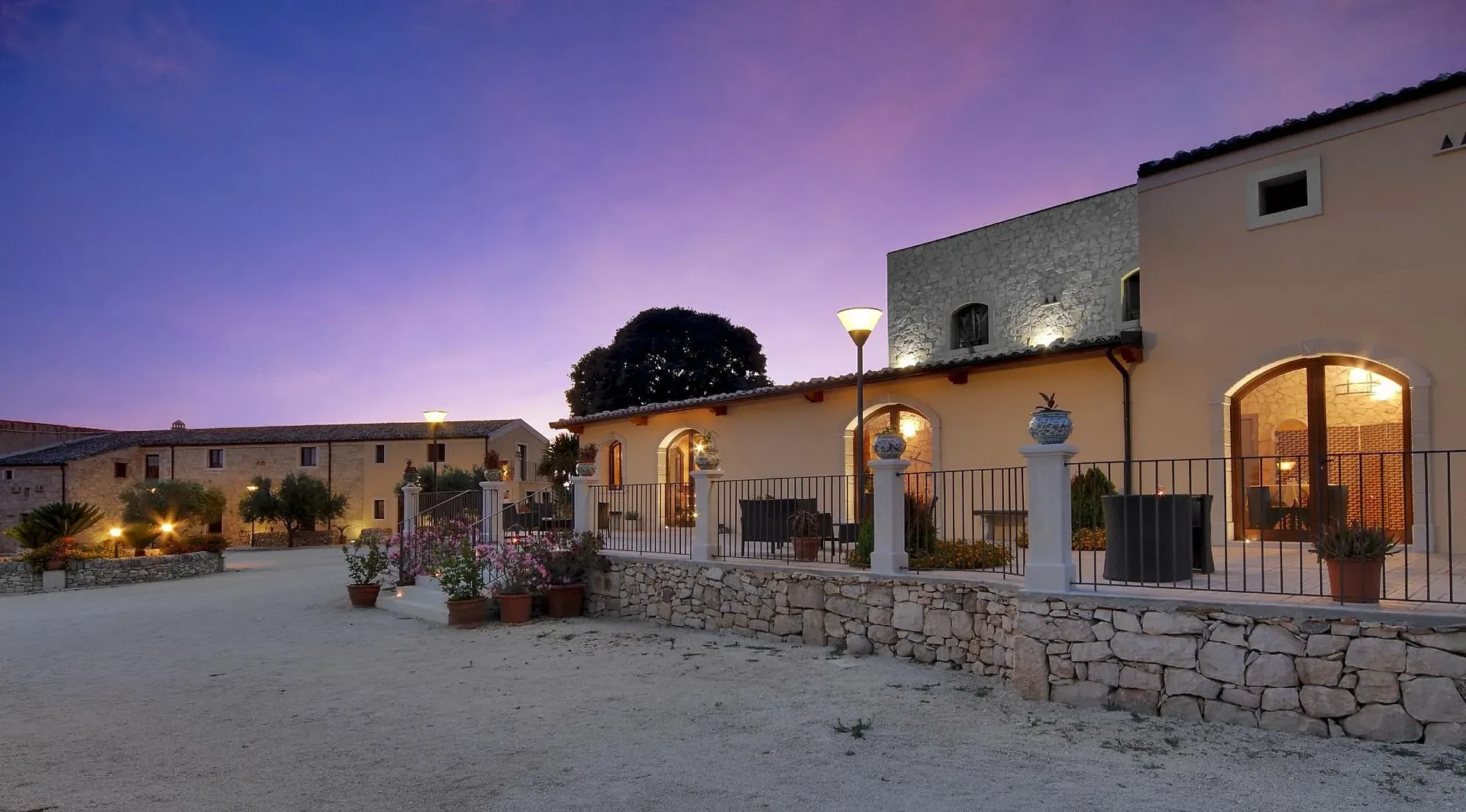 Esterno del resort in pietra al tramonto con cielo violaceo e cortile illuminato