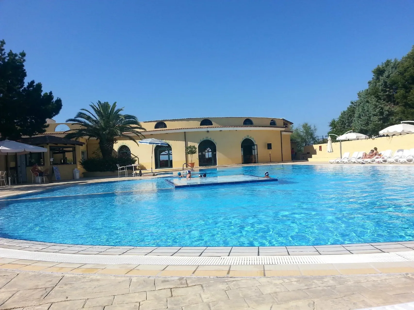 Piscina con bagnanti e edificio giallo del resort con palma e ombrelloni