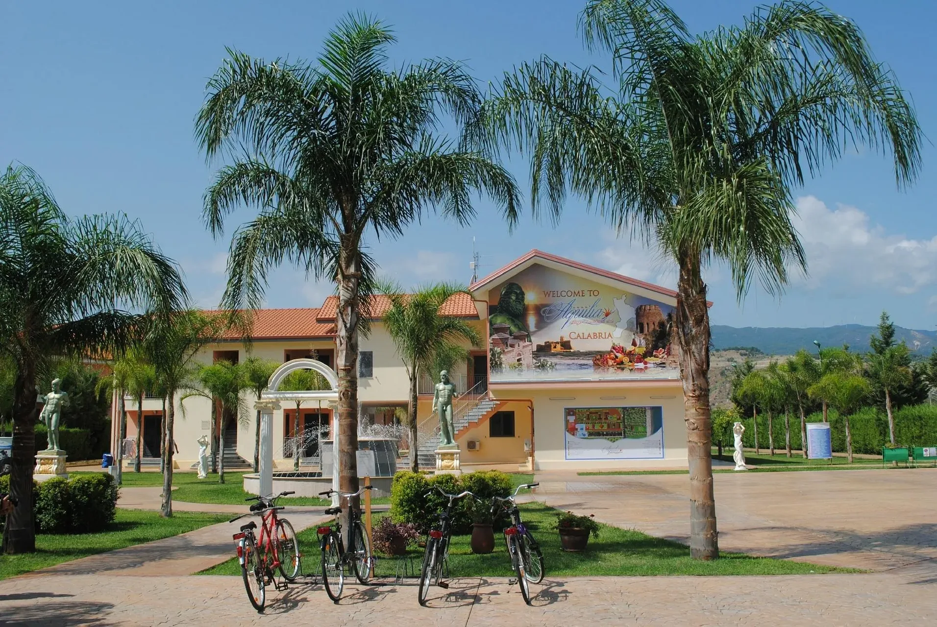 Ingresso del villaggio con palme, statue, fontana e insegna di benvenuto