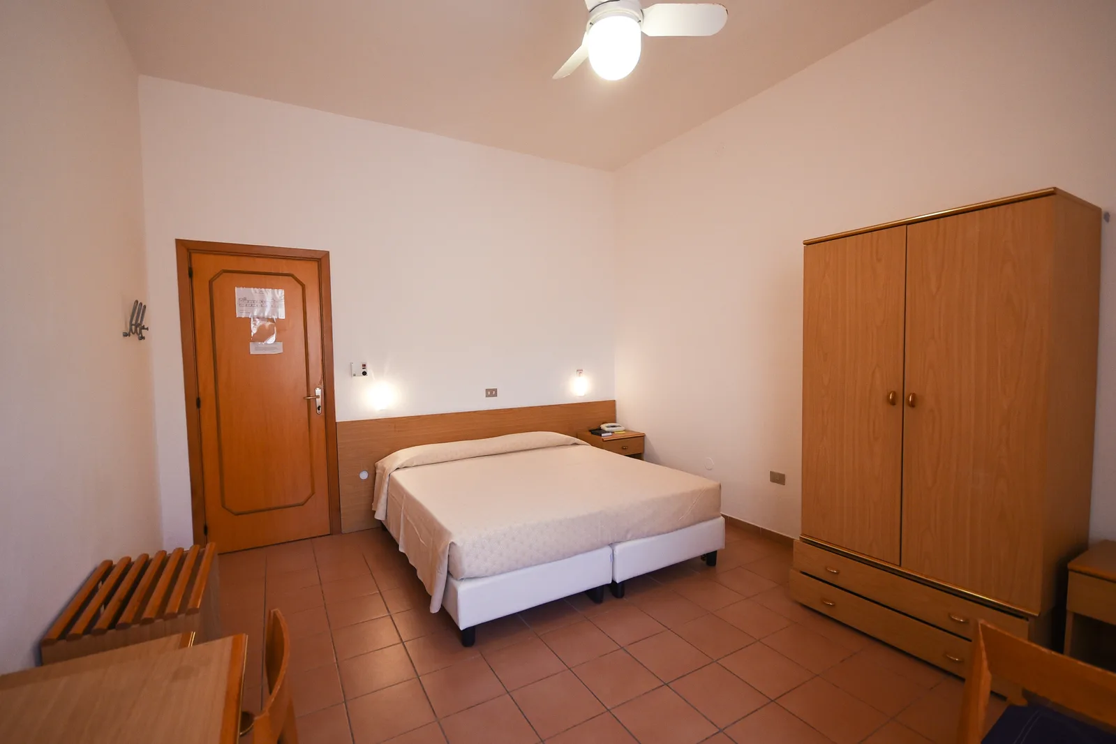 Camera matrimoniale essenziale con letto, armadio in legno, ventilatore a soffitto e pavimento in cotto