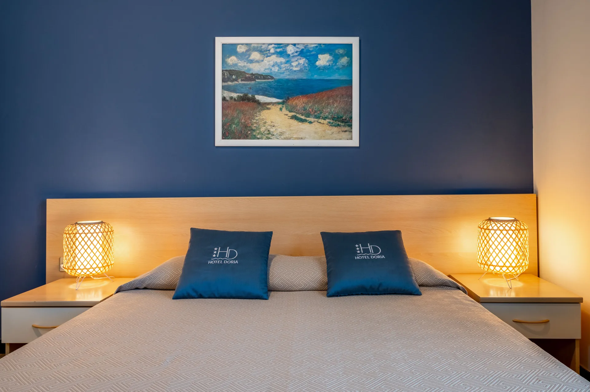 Testata letto in legno con parete blu, quadro marino e lampade in rattan accese