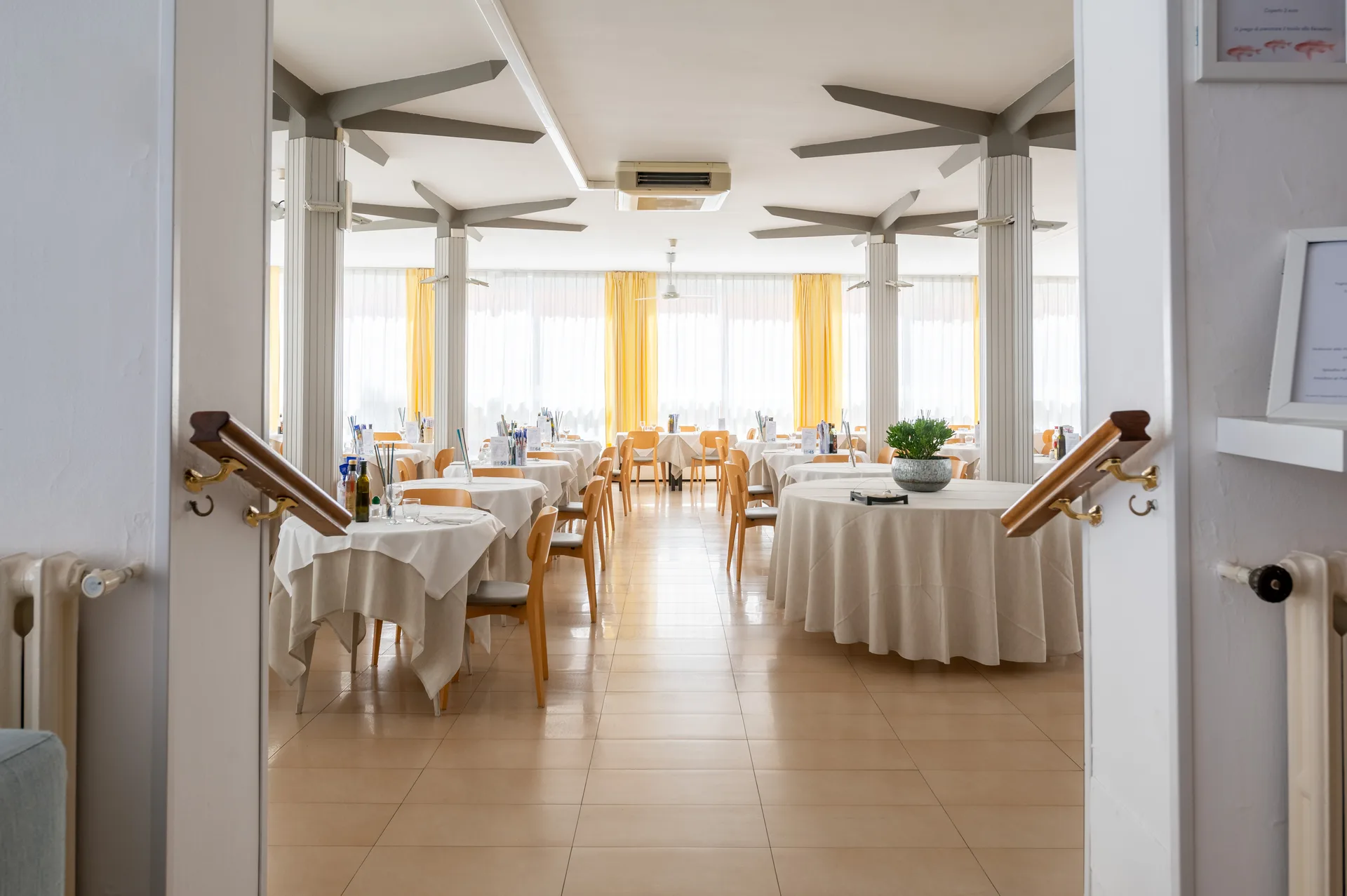 Sala ristorante luminosa con tavoli apparecchiati, tende gialle e colonne bianche