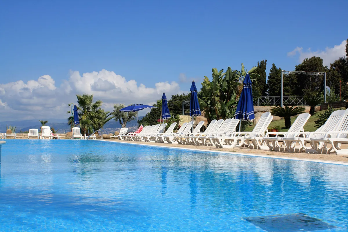 Piscina con lettini bianchi e ombrelloni blu circondata da palme e vegetazione