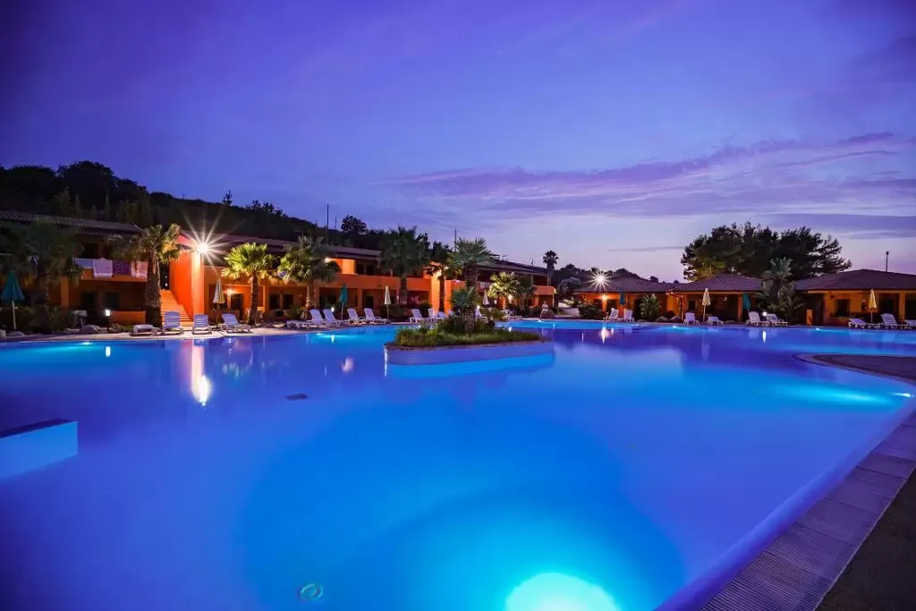 Piscina illuminata di blu al crepuscolo con villette arancioni e palme