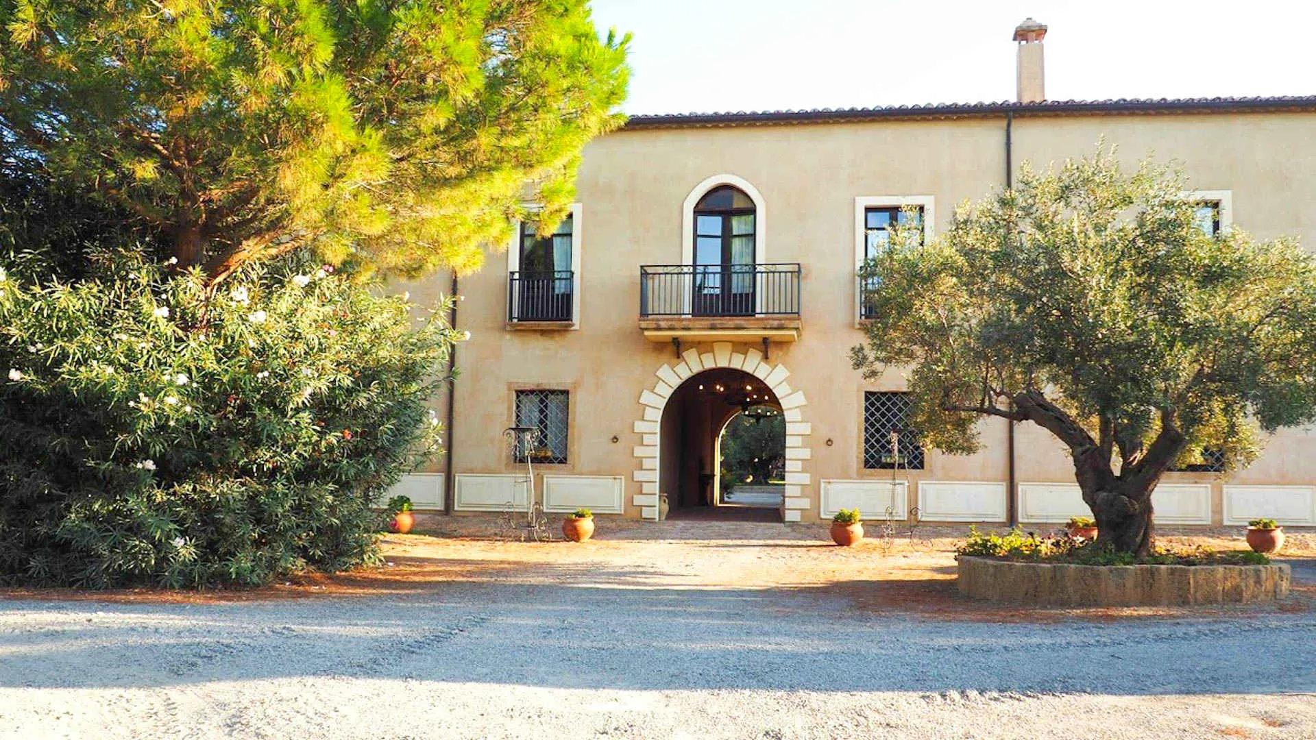Facciata del casolare storico con arco d'ingresso, balcone e ulivi nel cortile in ghiaia