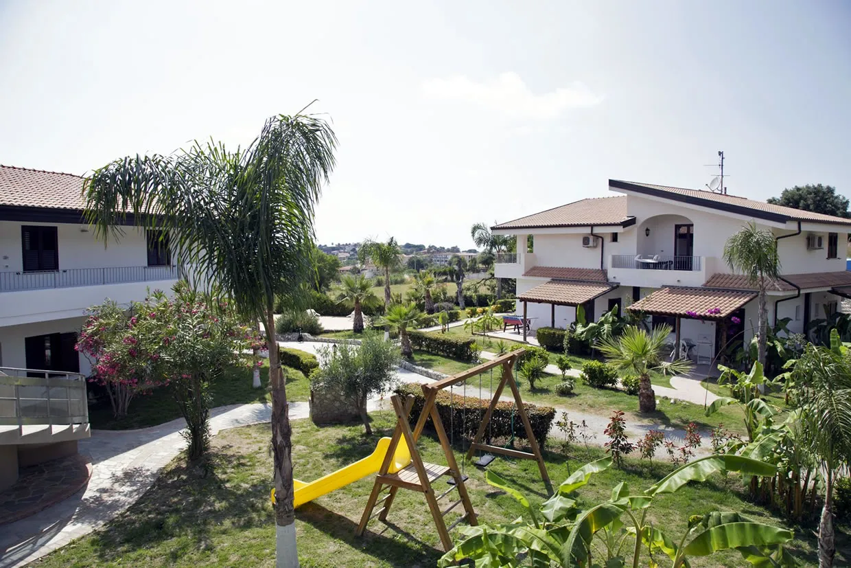 Giardino del residence con palme, area giochi per bambini e edifici bianchi