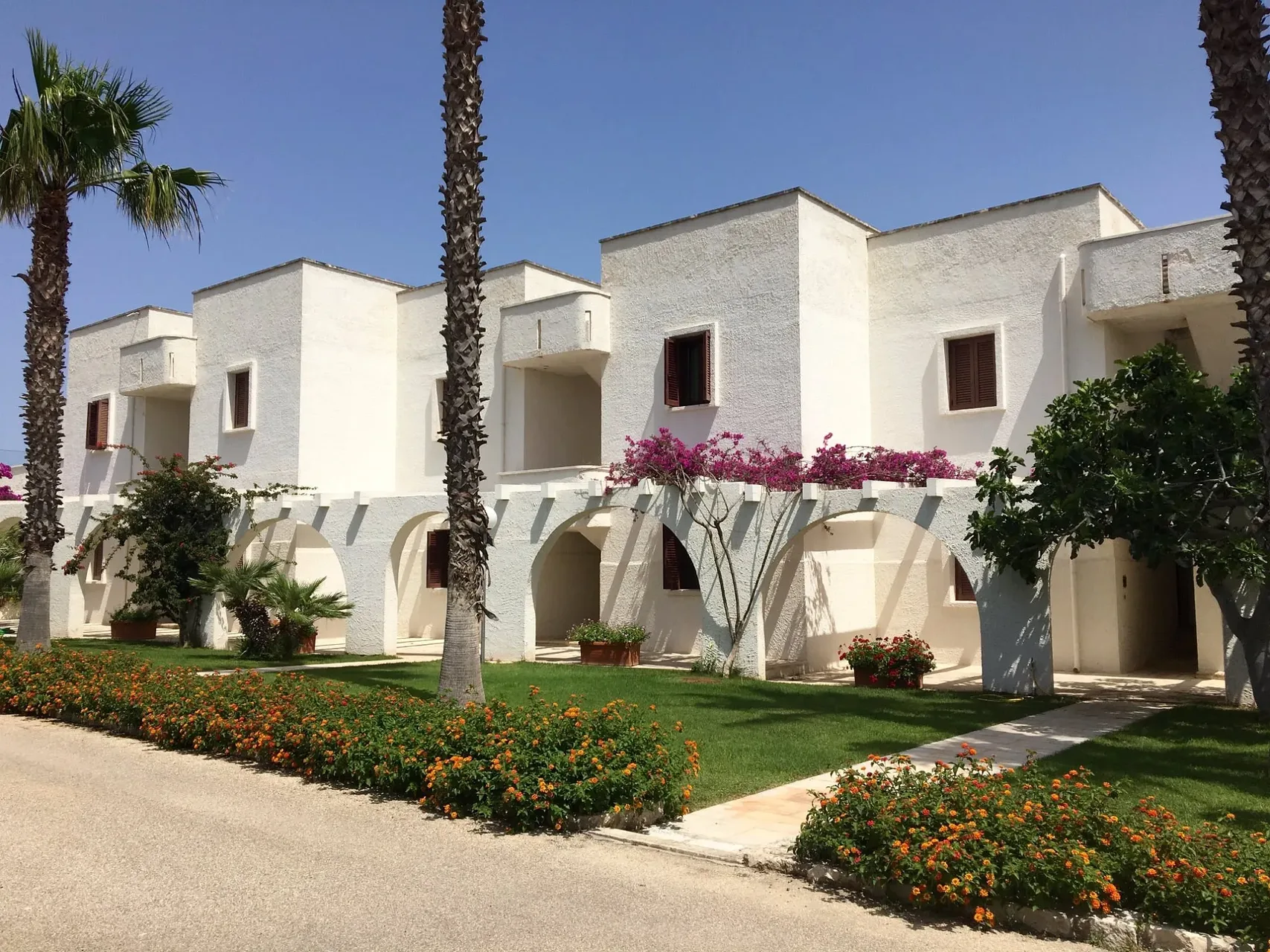 Edifici bianchi del residence con archi, palme e aiuole fiorite lungo il viale