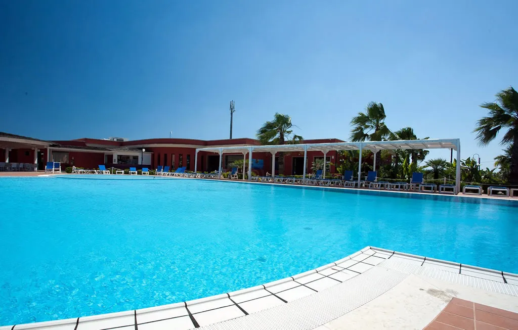 Ampia piscina del resort con lettini blu e pergolato bianco, edificio rosso sullo sfondo