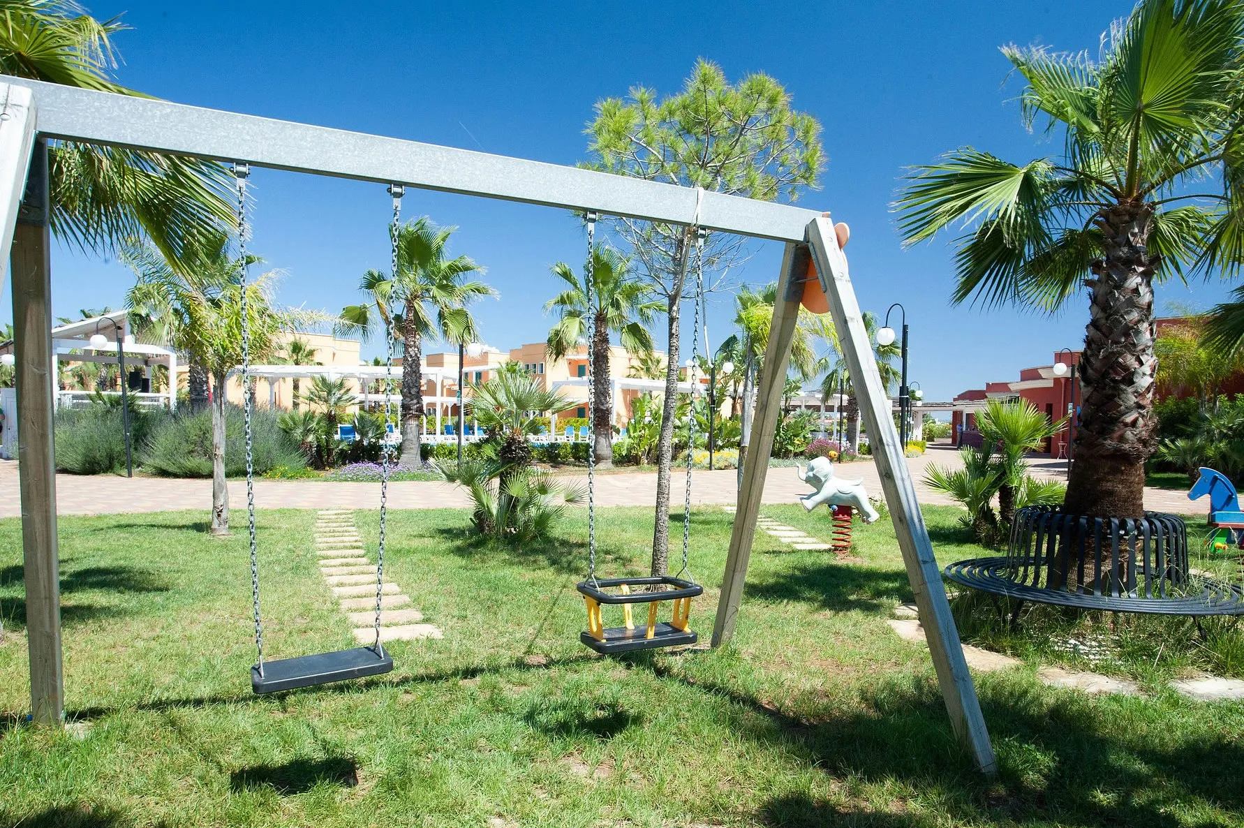 Area giochi per bambini con altalene e dondoli su prato verde, palme e residence sullo sfondo