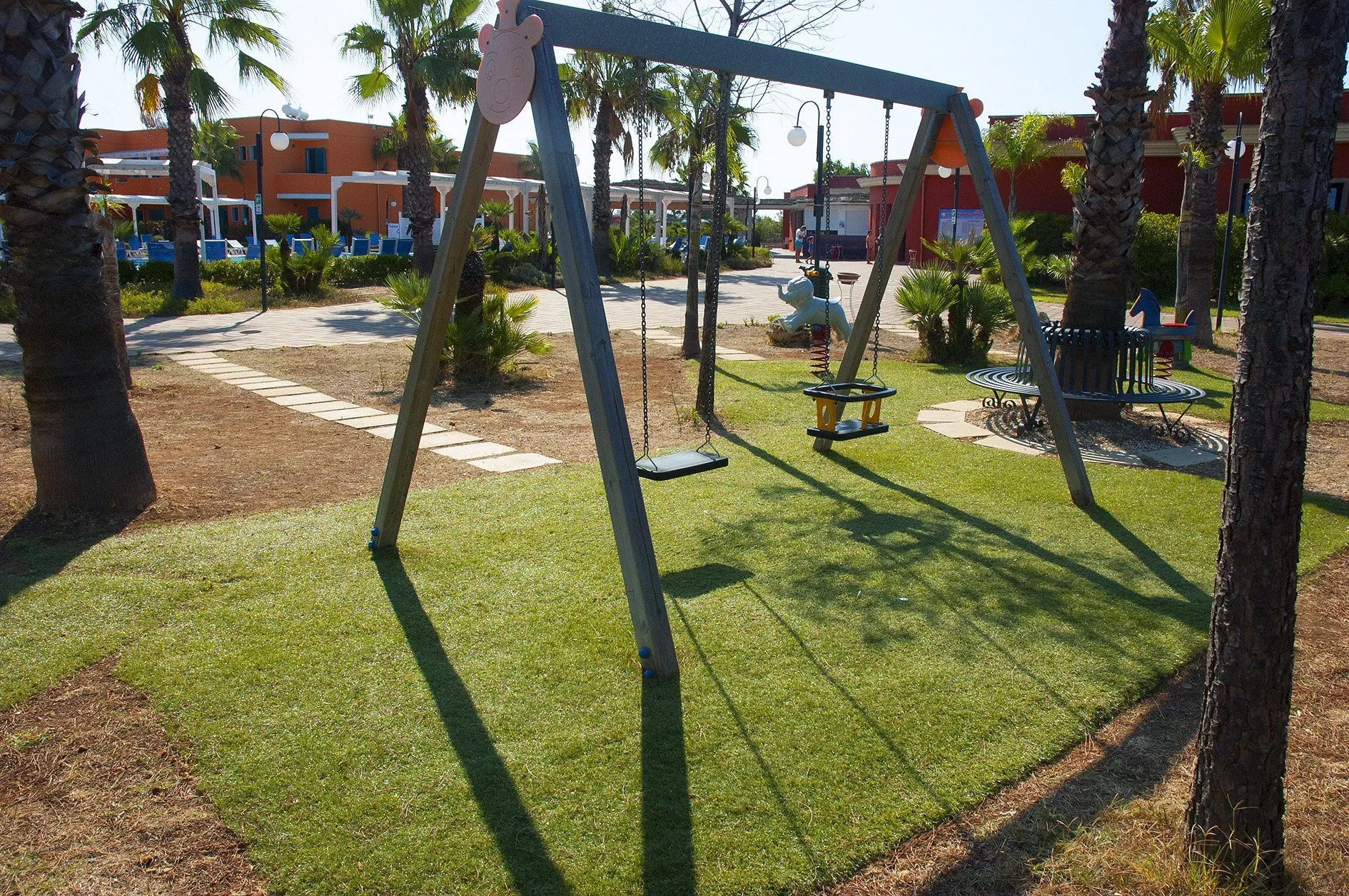 Parco giochi per bambini con altalene tra le palme, vialetto e edifici del resort sullo sfondo