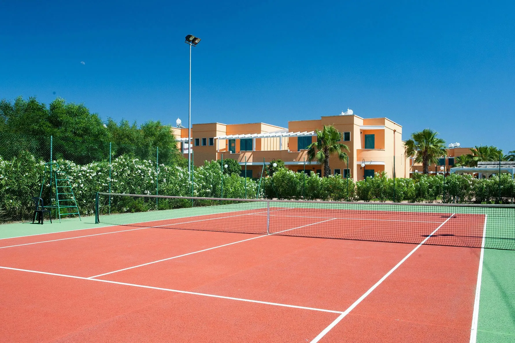 Campo da tennis in terra rossa con rete, circondato da siepi fiorite e residence color ocra
