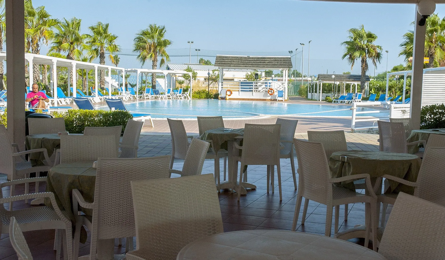Area bar con tavoli e sedie bianche affacciata sulla piscina circondata da palme