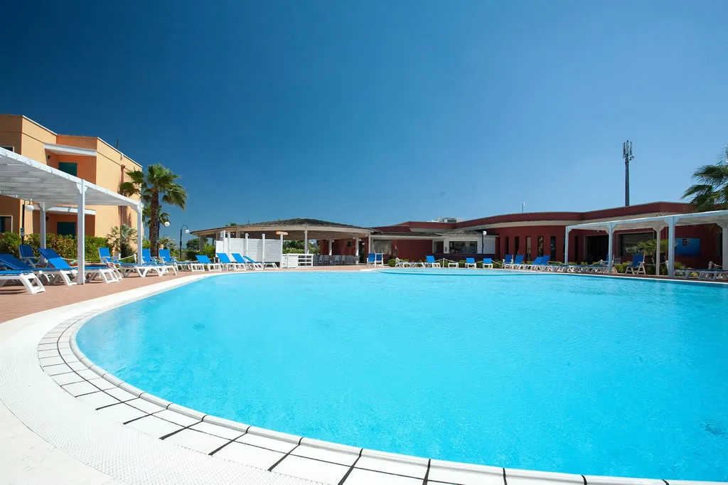 Piscina rotonda del resort con lettini blu e bianchi, edificio rosso e palme sullo sfondo