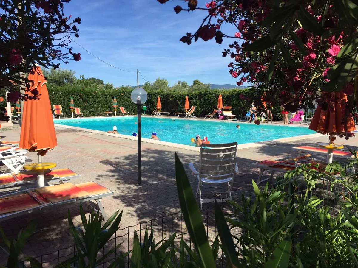 Piscina con bagnanti, lettini e ombrelloni arancioni tra fiori e vegetazione