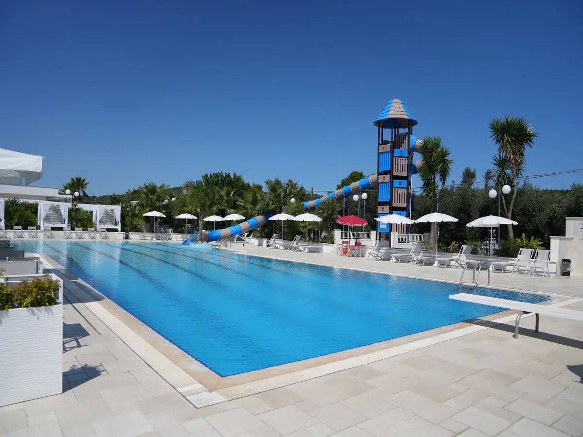 Ampia piscina rettangolare con scivolo a torre blu, lettini e ombrelloni bianchi sul bordo