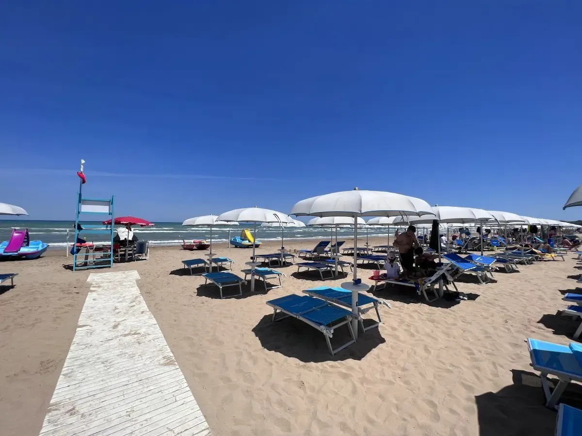 Spiaggia con ombrelloni bianchi, lettini blu e bagnanti, mare e cielo azzurro sullo sfondo