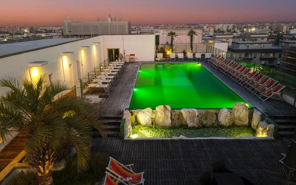 Piscina sul tetto illuminata di verde al tramonto con lettini arancioni e vista sulla città