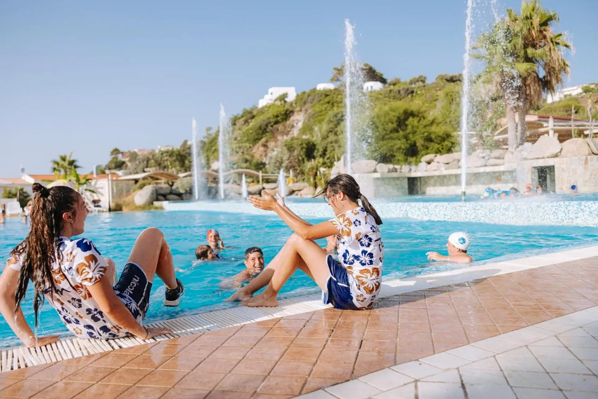 Ragazzi che giocano a bordo piscina con giochi d'acqua e fontane in una giornata di sole