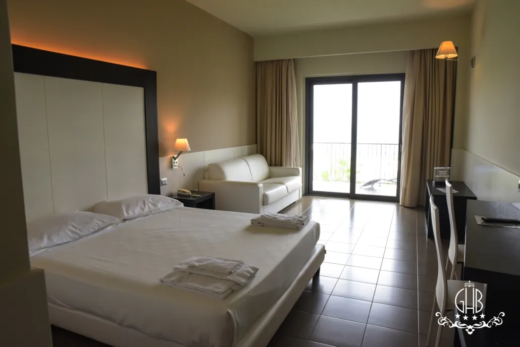 Camera spaziosa con letto matrimoniale, divano bianco e balcone con vista mare