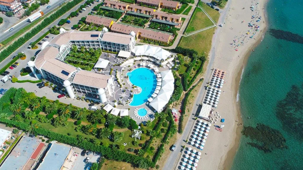 Vista aerea dall'alto dell'hotel con piscina a forma sinuosa, giardini e spiaggia con ombrelloni