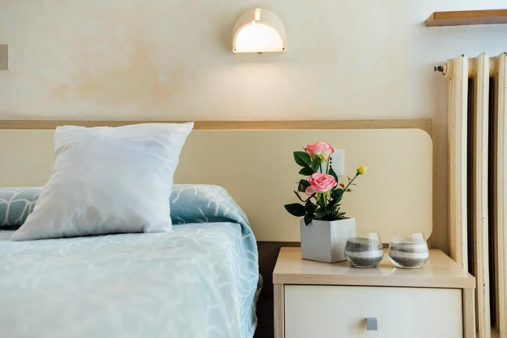 Dettaglio del letto con cuscino bianco, comodino con fiori rosa e applique illuminata