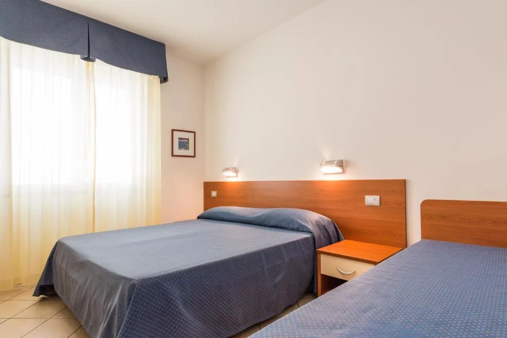 Camera doppia con letto matrimoniale e letto singolo, testiera in legno e finestra con tende chiare