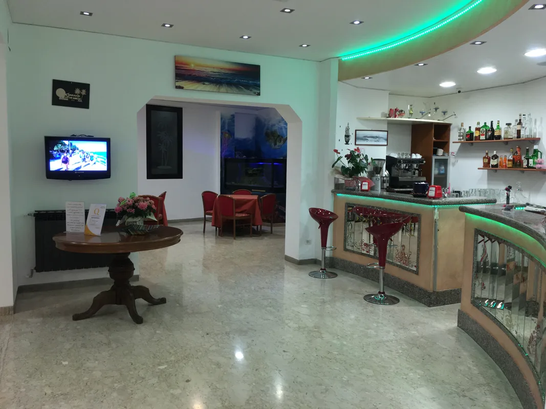 Hall dell'hotel con bancone bar, sgabelli rossi, tavolo in legno e zona soggiorno con TV