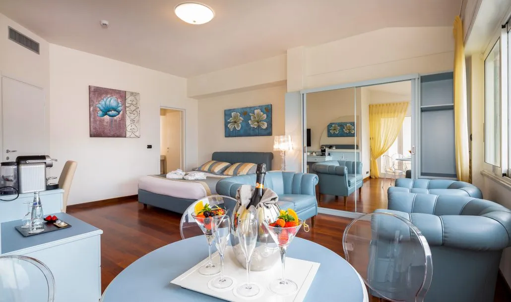 Ampia suite con letto, poltrone blu e tavolino con champagne e frutta di benvenuto