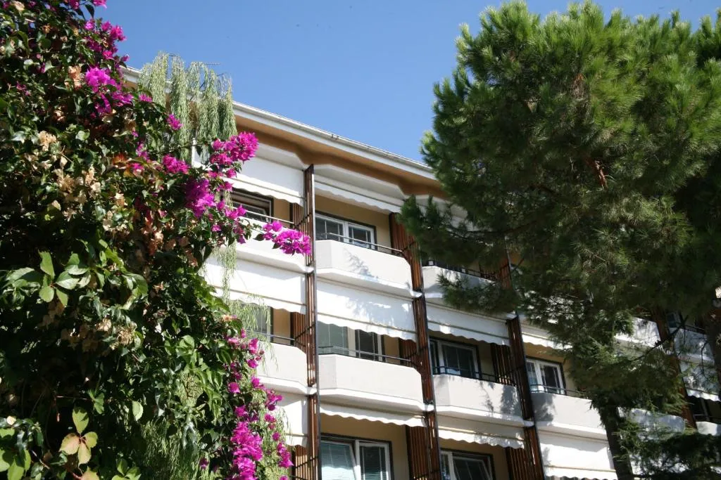 Facciata dell'hotel con balconi, circondata da pini e fioriture di bouganville