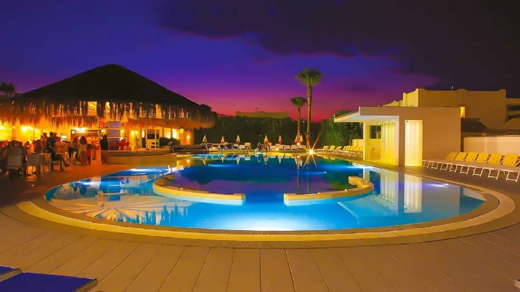 Piscina illuminata al tramonto con cielo viola, bar in paglia e ospiti durante la serata