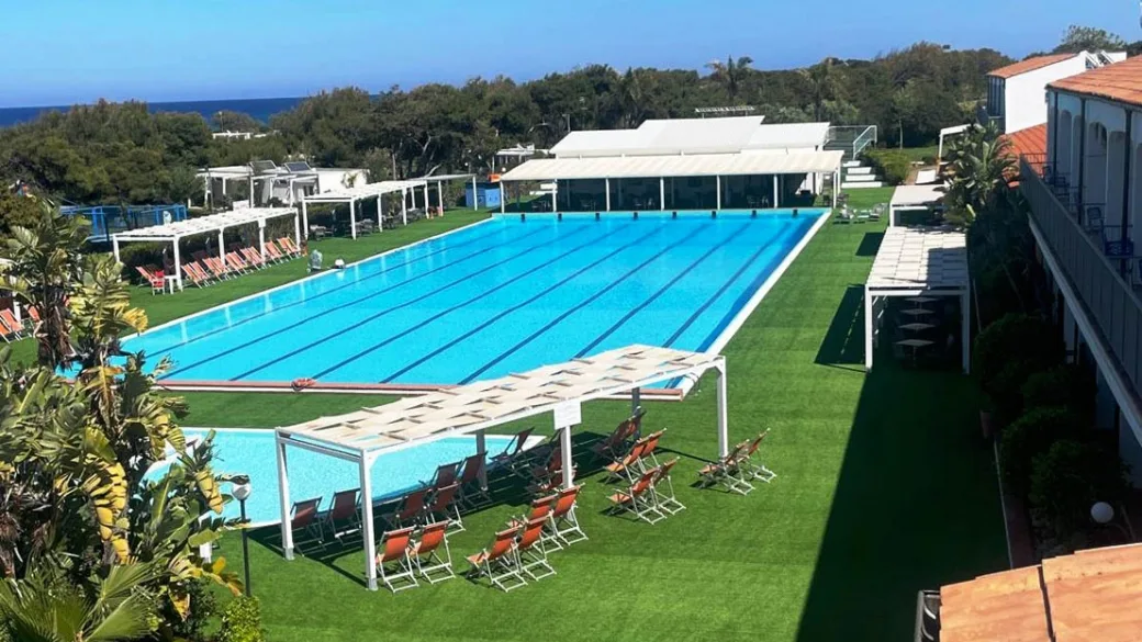 Piscina olimpionica con prato verde, gazebo bianchi e lettini arancioni, vista mare sullo sfondo