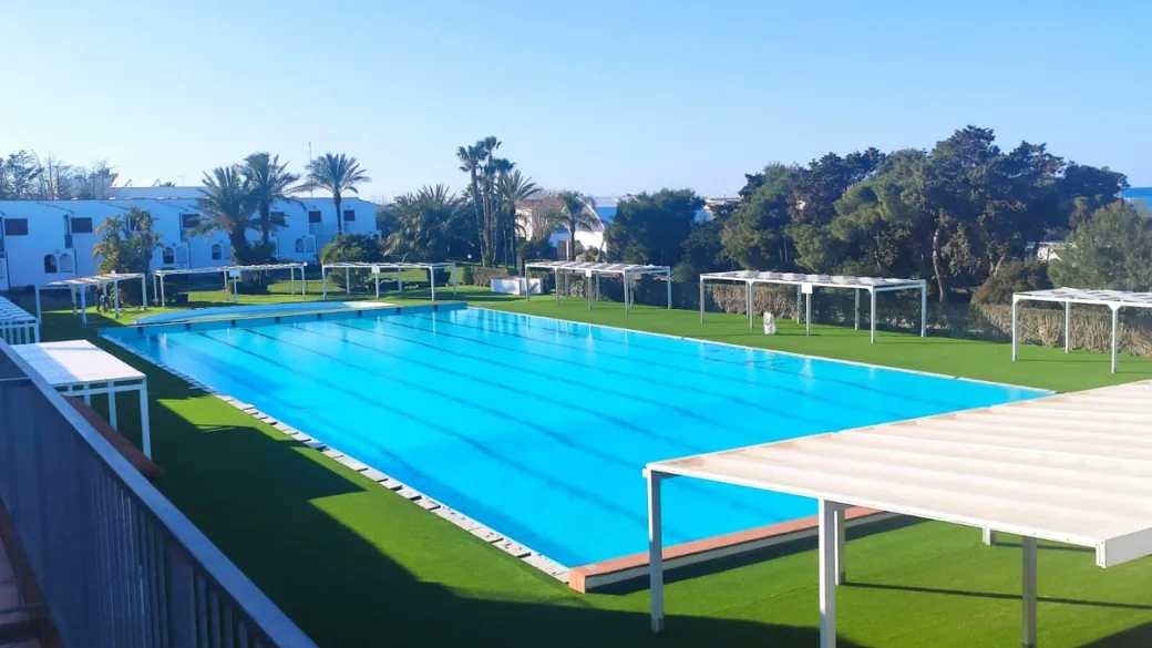 Piscina a corsie circondata da prato verde, gazebo bianchi e alberi con mare all'orizzonte