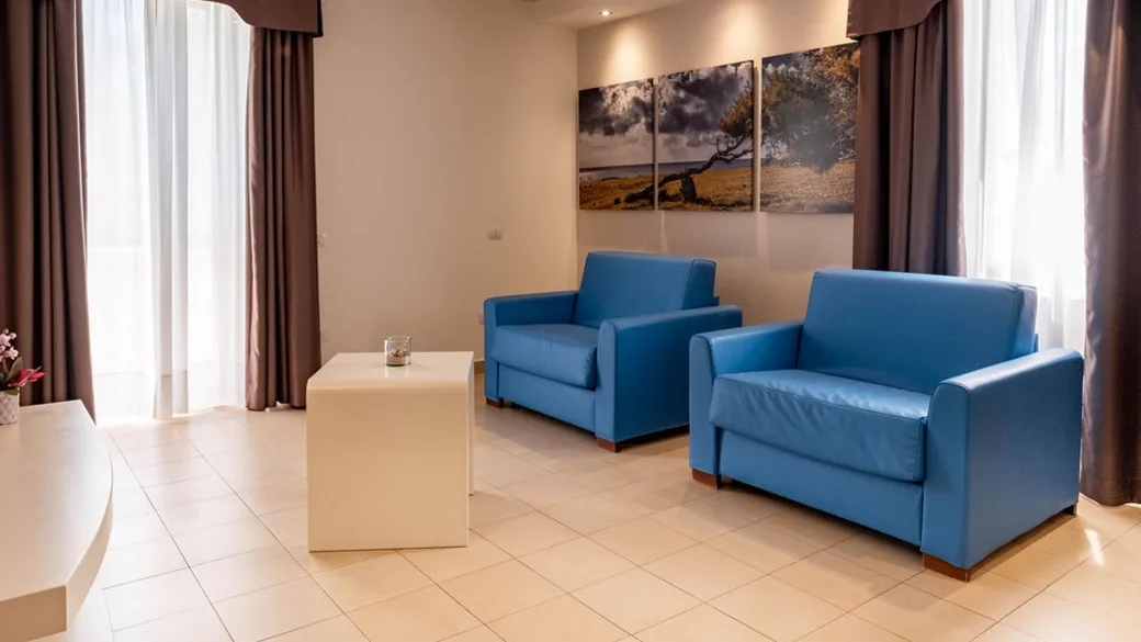 Zona soggiorno di camera con due poltrone azzurre, tavolino e quadri decorativi alle pareti