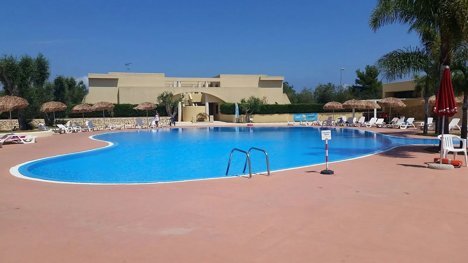 Ampia piscina con pavimentazione rossa, ombrelloni in paglia e edificio del villaggio sullo sfondo