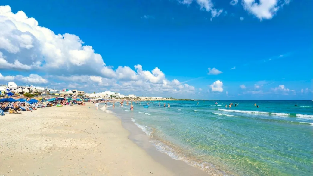 Spiaggia sabbiosa con mare cristallino, ombrelloni e bagnanti sotto un cielo con nuvole