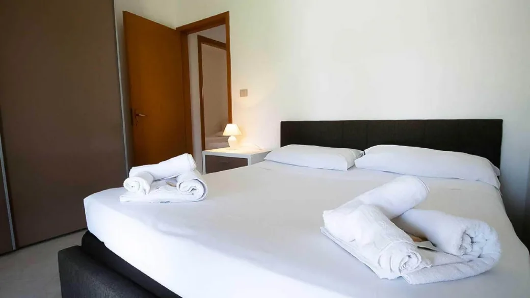 Camera da letto matrimoniale con lenzuola bianche, asciugamani arrotolati e comodino illuminato
