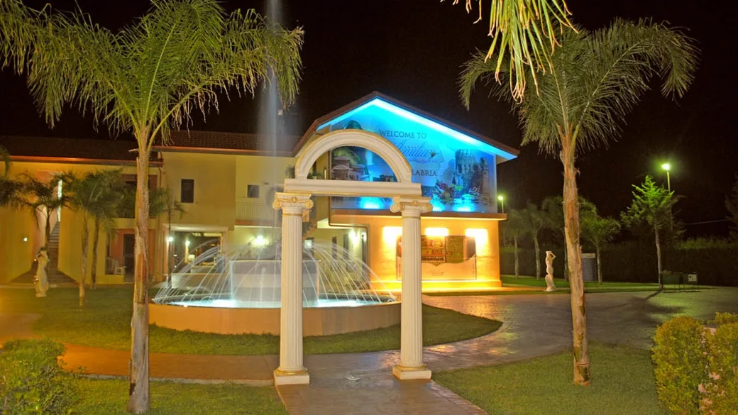 Ingresso del villaggio illuminato di notte con fontana e arco classico