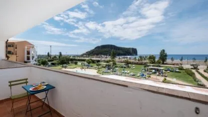 Balcone con vista mare, piscina, area verde e spiaggia con sdraio, sotto un cielo azzurro con nuvole.
