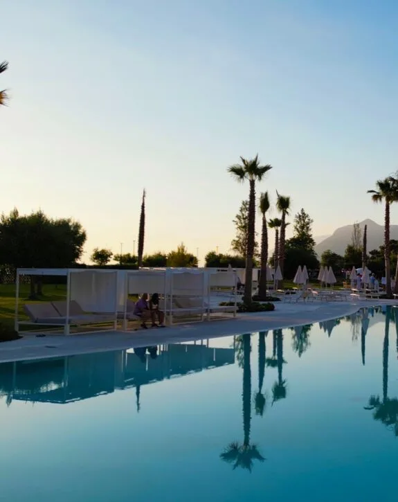 Piscina con lettini e palme, al tramonto con montagne sullo sfondo.