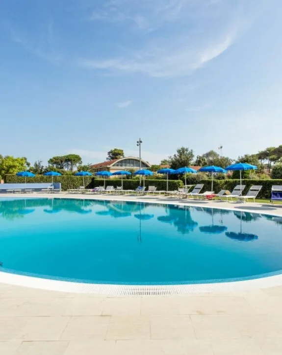 Piscina rotonda all'aperto con ombrelloni blu e lettini, cielo sereno e vegetazione sullo sfondo.